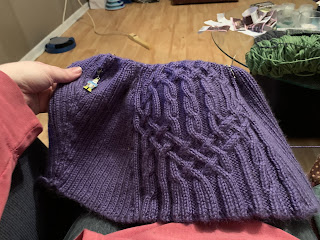 In progress: the lower body of a sweater knit in the round featuring a wide intricate cable panel and slanted 2 by 2 ribbing on either side of it. The yarn is a luscious purple and the silver stitch markers on the circular needle appear as little glints against the rich background color.