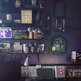 Black shelves with ornate (but difficult to see) brackets upon which are art materials; skull tub with purple and green dragon-scale paint brushes, vintage tins, ivory drawers, a purple box with silver strapping and latches. Next to the shelves are a variety of Gothic arch candle holders. On the left is a tall, slim dark brown one with gold trim and a mirror, it is also a sconce for a purple dinner-style candle. Central at the top is a black metal arch with some swirls and three candle holders, but the candle-holders are being used to suspend three purple glass hearts. Bottom centre is a dark brown wooden Gothic arch, short and squat, with three metal wire Gothic arches inside it, a mirror and a Highlands & Islands Scottish Green Party sticker. On the right is a crescent moon mirror of black metal, also with three candle-holders. The wall they are on is dark purple. To the right there is a black metal shelf with trays of pencils and charcoals. The computer monitor is on the left. In the middle of the desk are a grey vintage-style desk-lamp, a computer tablet in a black metal recipe book holder, and what looks like a gold and purple antique book but is actually a Harry Potter book lamp. The desk is black marble effect. Behind the monitor, lamps, tablet and bottom of the metal shelves is greyscale Gothic architecture patterned wallpaper under a black dado rail. You are very patient listening to this description, thank-you.