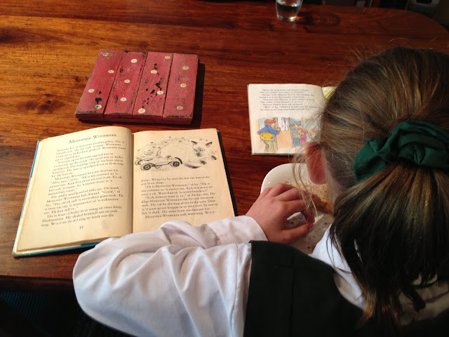 Anna Sophia eating lunch at the kitchen table in her school uniform while reading a book with another at the ready.