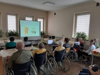 Na tle sali ośrodka, której ściany mają kolor beżowy widzimy siedzących pensjonariuszy - czytelników, którzy oglądają prezentację  i słuchają opowiadań pani bibliotekarki na temat historii książki i bibliotek