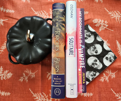 The spines of three books - Teach the Torches to Burn, Solitare, My Roomate is a Vampire - face up. Bookended by a black staub ceramic pumpkin witha gold stem handle tot he left and black and white skull napkins to the right. Orange with white leaf pattern in the background