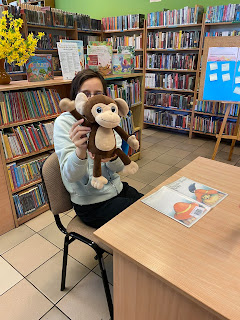 Pani bibliotekarka z zielonej bluzie siedzi przy jasno beżowym stoliku i pokazuje do aparatu pluszową małpkę. W tle widać ladę biblioteczną i półką na której stoi żółta forsycja w brązowym wazonie.