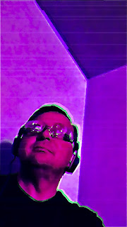 a freak wearing headphones and mad scientist glasses is glowing  with a green light as he cranes  his neck upwards in a purple room.