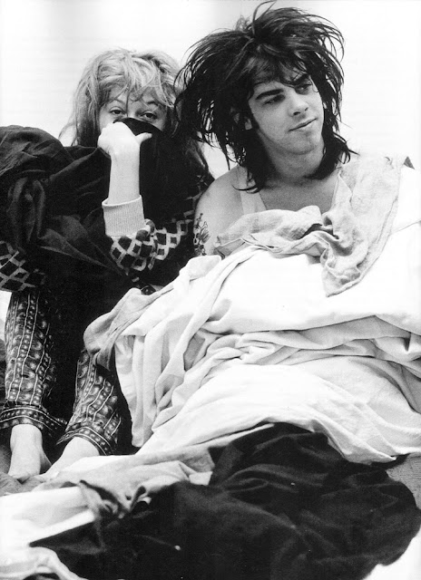 Nick Cave looking off camera right while laying down leaning against Anita Lane who is sitting with knees up and partially covering her nose and mouth and looking directly at the camera