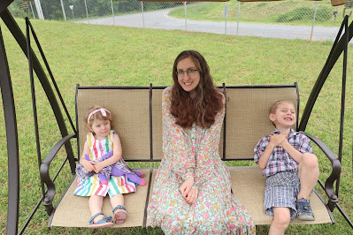 a smiling woman in a flower covered long dress sits on a swing with two small children