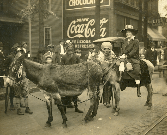Set in 1910. A woman in riding boots wearing a long coat and a wide brimmed hat rides a top of a mule. There are people in the background, almost all wearing hats, walking about. Next to the woman is a man in a clunky skeleton outfit and a man in completely black outfit with his face covered.