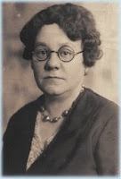 A B&W portrait of a middle-aged, plump white woman with round eyeglasses and short, curly, dark hair.