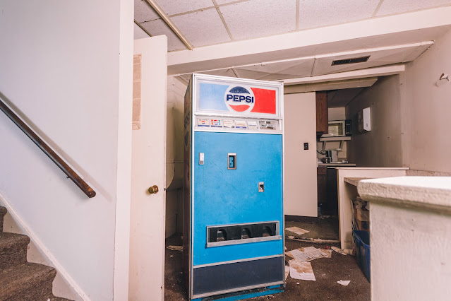 An old Pepsi vending machine stands abandoned in a neglected room with torn papers on the floor and a stairway leading to an unkempt office area, reflecting a sense of disuse and forgotten daily routines in a once bustling radio station.