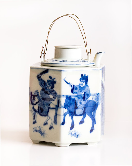 A Japanese teapot in blue and white design with horses and riders, and a wire handle.