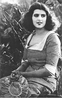 A young woman with thick, dark, shoulder-length hair sitting outdoors. She is wearing a dress with a fine checked pattern and holding a matching hat.