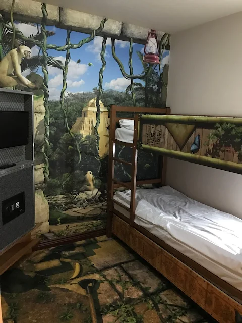 A television and safe are on the left, a bunk bed on the right and mural carpet and wallpaper showing crumbling aztec temple, vines and a monkey