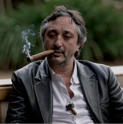 Harmony sitting outside wearing a black leather blazer and smoking a cigar on the chest up