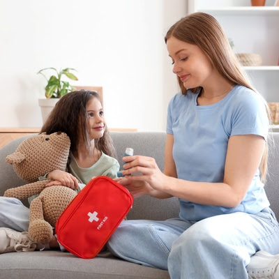 A woman sits on a sofa with a young girl who holds a teddy bear. The woman holds an inhaler and a first aid kit.