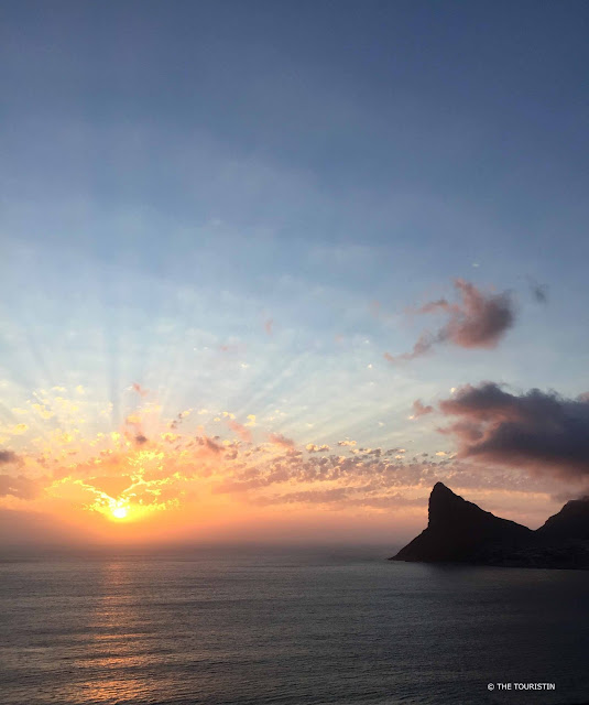 Sunset over the ocean next to a pointed hill on shore under a fluffy cloud.. 