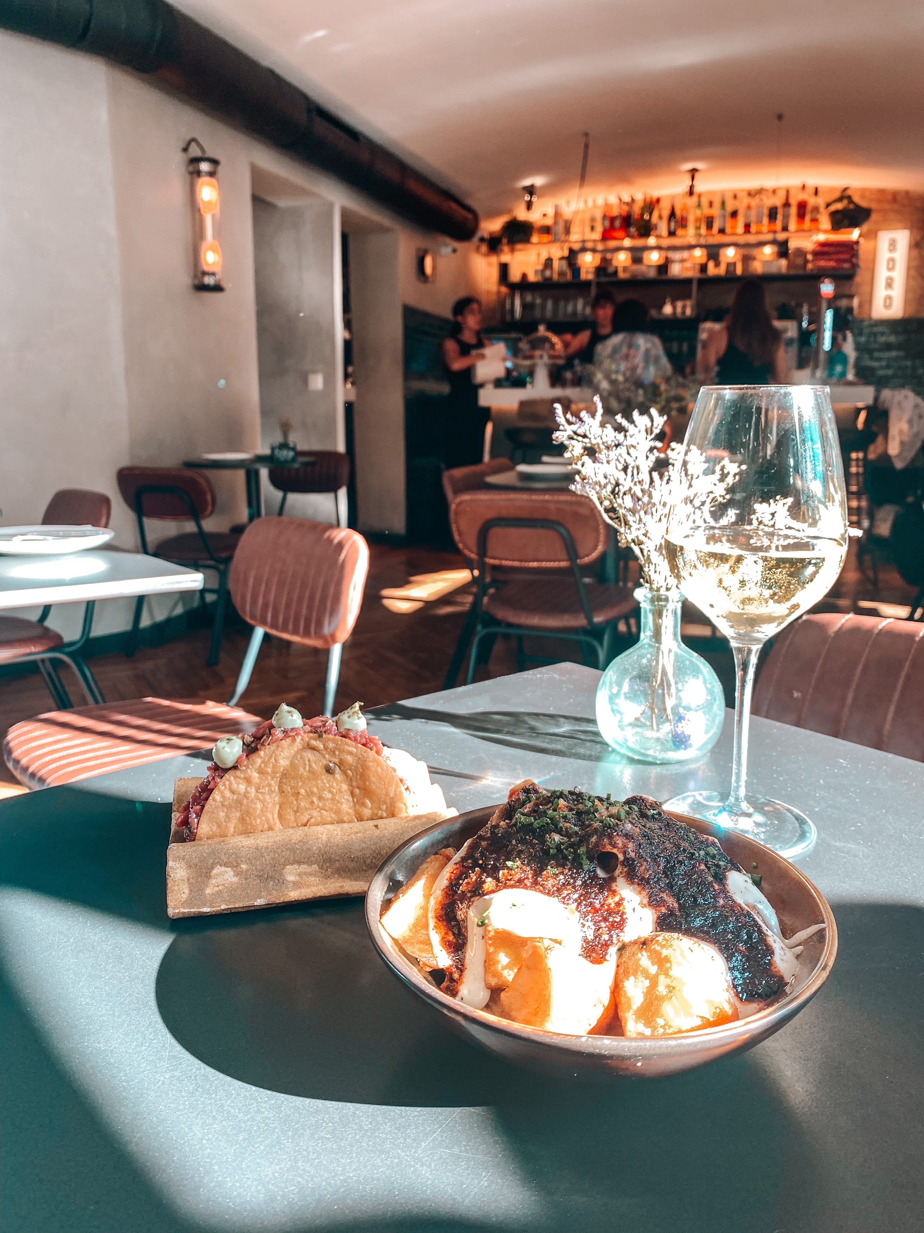 tables dans le restaurant boro à Barcelone avec dessus un verre de vin blanc des patatas bravas un tacos et un bouquet de fleurs séchées dans un vase bleu transparent et dans au fond le comptoir avec deux serveuses et deux clientes