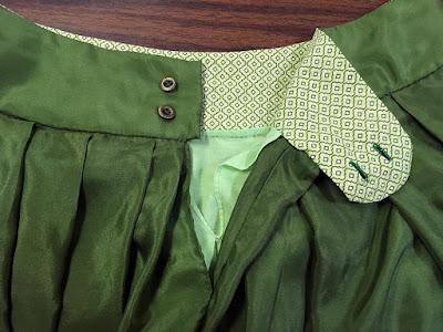 The waistband of a pleated green silk skirt, with the buttonhole tab folded back to show the patterend green lining, two neat horizontal buttonholes, and two brass buttons.