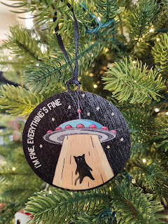 small wood circle shaped ornament with a cat being beamed up to a space ship In white print, along the edges it says I'm fine, everything is fine