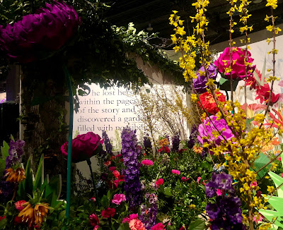 A garden of pink, yellow, and purple flowers with a page from a book as the background wall.  On the page, it says, "She lost herself within the pages of the story and discovered a garden filled with possibilities."