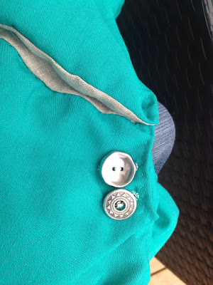 A pair of buttons on a bright turquoise skirt with a grey-lined pocket. One button is plain, slightly dished silver with two holes, and the other is a disc with an opening in the middle and a bar, and a stamped sun ray design around the rim.