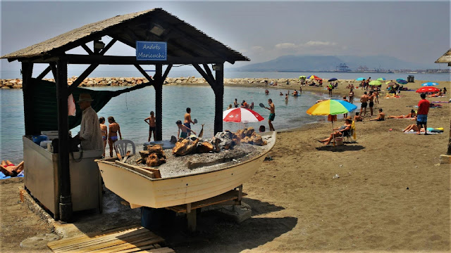 Barca de espetos y su quiosco de madera en una playa llana de arena con mucha gente y el agua del mar al fondo.