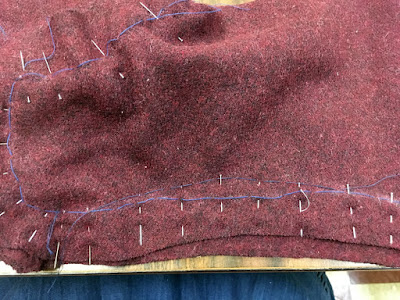 A close-up of the garnet-red hose back seam at the ankle, showing the pin heads a fraction of an inch further in than the white chalked line and blue basting stitches, with a new set of basting stitches tracing the lines of the pin heads.