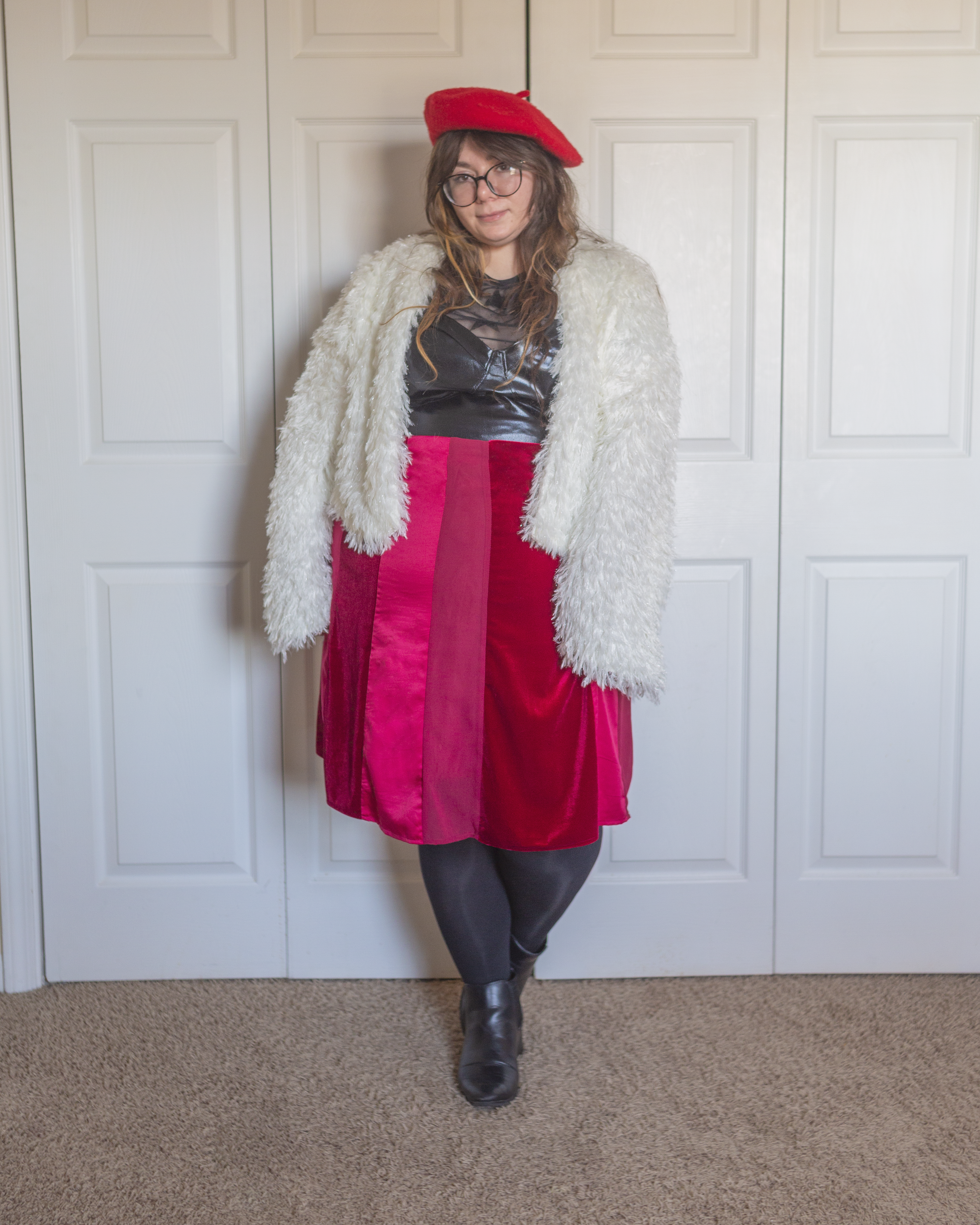 An outfit consisting of a reed wool beret, a white faux fur jacket over a black sheer star print top with a leather camisole over, tucked into a dark pink, light pink, and berry cut and sew style a-line midi skirt and black Chelsea boots.