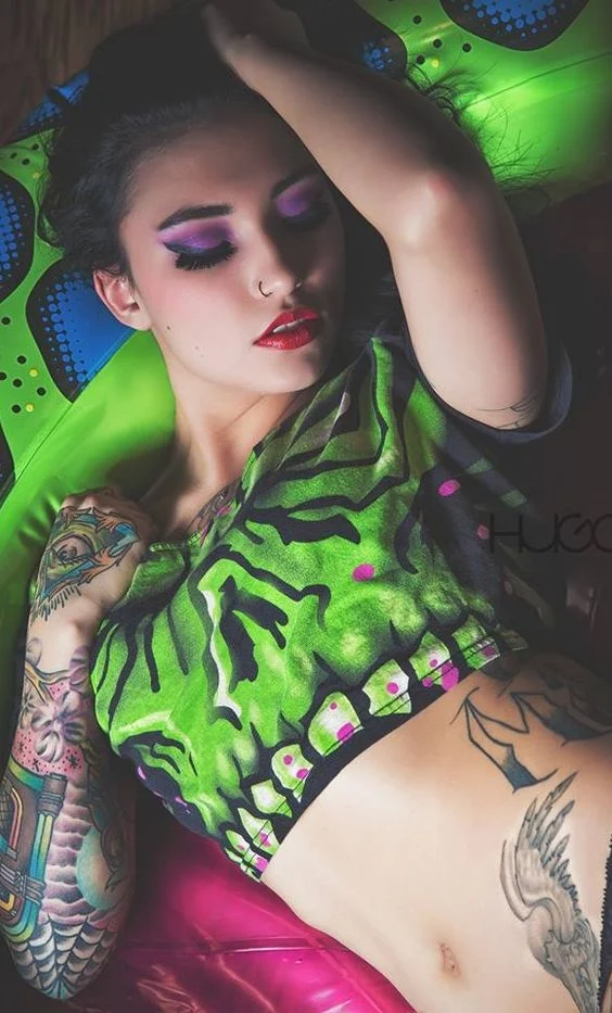 Vemos a una mujer tendida en un colchon hinchable verde, la chica lleva tatuajes en el antebrazo de una gramola, y tattoo de alas en la cadera
