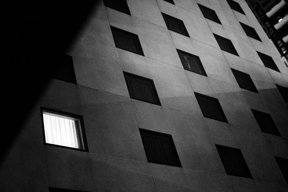 Black and white photo taken at night-time of a hotel building with all the square bedroom windows dark, except one. Taken by Jim Campbell, in Melbourne