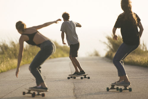 Fotografia de três skatistas, um homem ao centro e mais a frente e duas mulheres, uma em cada lado, andando de skate em uma estrada. Eles estão de costas, andando em direção ao fim da estrada.