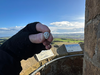 A photo of a small ceramic skull (Skulferatu 118) being held up with the view from Hopetoun Monument in the background.  Photo by Kevin Nosferatu for the Skulferatu Project.