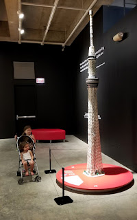 Two young girls with a stroller stand next to a large LEGO tower.