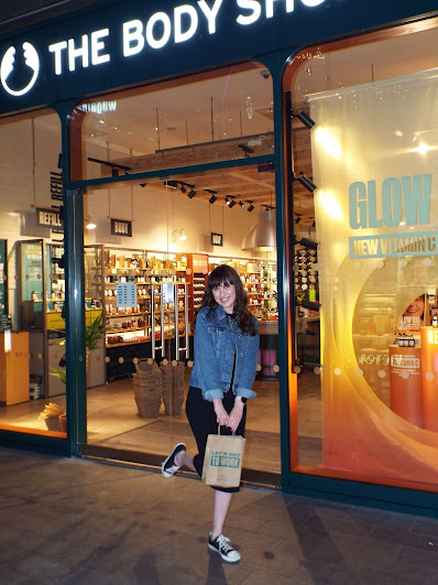 Blogger, Megan Goodier, posing outside the warmly lit body Shop Liverpool One, with leg popped up and a very sweet smile!