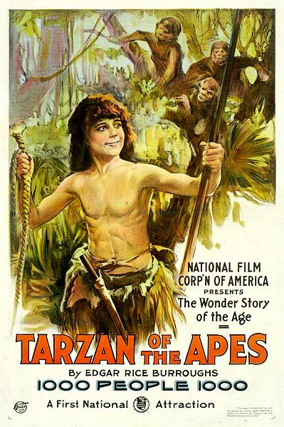 a bare-chested smiling boy is wearing a grass skirt and holding a rope in one hand and a pole in the other, as a group of monkeys looks on from the tree behind him