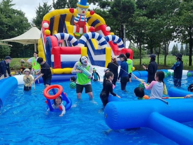 alt="계양아라온 워터축제에서 어린이들이 물총놀이를 즐기고 있는 물놀이존 풍경. 중앙에 광대 복장을 한 스태프가 아이들과 함께 놀고 있으며, 뒤에는 대형 에어바운스 슬라이드가 설치되어 있음"