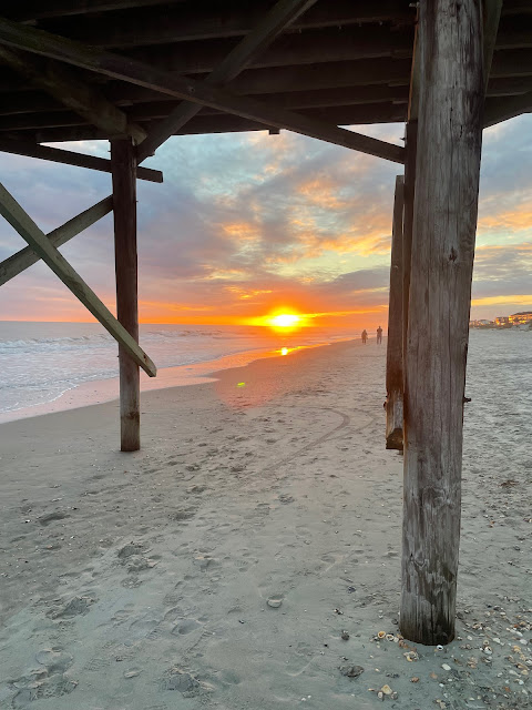 a view of the sunset from underneath the pier. the sky is mostly obscured by the pier, but the glow of the sun is visible. The beach is empty.