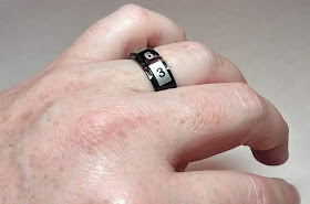 A closeup of the author's left hand, showing a black metal ring on the ring finger. The ring is made of two bands, an inner band which sits against the finger, and the outer band, which spins freely around the inner band. The outer band is painted in alternating blocks of black and grey, with a different number printed in each block. There are two small arrows on the inner band to indicate which number is the result of the 'roll.'