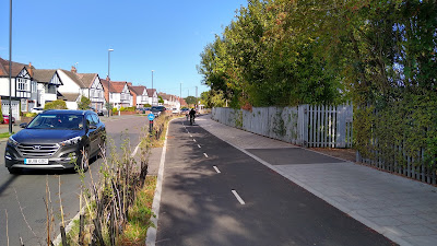 A two way cycle track on the right side of a street with houses far left. The track is protected by a recently planted hedge. There is a grey footeay to the right.