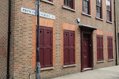 Streetcorner and part of a street; yellow brick row house, formerly likely a weaver's house and workplace, with heavy red-painted wooden shutters and door at ground level.  Red-painted window frames and glass windows at second floor level. Sign reading Princelet Street E1. Lamp pole.