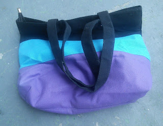 A bag striped in Purple, Turquoise, and Black.  It has two handles and a zippered top. 