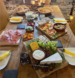A table with platters of food on top. There's a cheese board with grapes and crackers. There's a meat board with charcuterie. There's a fish board with prawns, salmon, mackerel pate and chunks of lemon, and there are salad items dotted about.