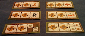 Six construction tiles. Each is long and narrow with room for three icons in a row. All of them have two icons representing a primitive stone house in the middle of construction. The third icon is different on all of them. Four tiles have a single icon in the third position: one shows a stone pillar with two feathers dangling from the capstone. This represents a shrine. Another has a stylized cartoon creature representing cattle. Another has a cartoon man with a beard and moustache in black clothes and headdress holding a staff, representing the shaman. The fourth three cartoon people in orange clothing, representing villagers. The last two tiles have multiple icons in the third space. One has two icons: a clay oven and a ring-shaped fence creating an enclosure. The final tile has four icons in the last space: the fence, the shrine, the oven, and the half-built house.