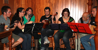 A group of people sitting behind music stands, holding ukuleles, discussing something animatedly.