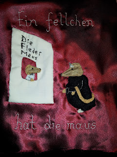 3a. The completed page, colours are slightly off because the fabric is so shiny it's hard to photograph. Text reads "Ein Fellchen hat die maus" - a furcoat has the mouse