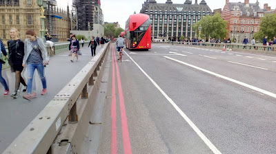 A view along Westminster Bridge. There is the houses of Parliament to the left in the background. On the bridge there is a footway to the left, a steel barrier to its right, then a painted cycle lane with a cyclists and then a bus lane with a bus in it.
