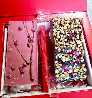 Two rectangular chocolate bars. One pink with long green thin branches on it and red blobs to look like cherry blossoms on it and one brown with with punk and green bits all over it on a bright background 