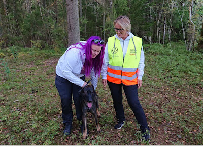 Linnéa Awwö Andersson och Susanna Johansson med hunden Herax framför sig i skogsmiljö.