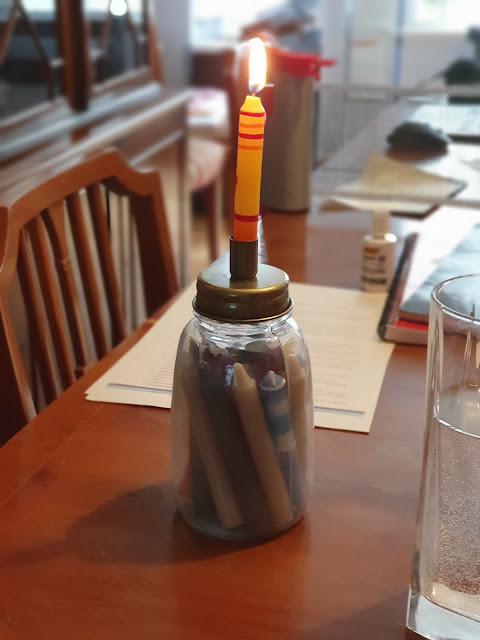 A candle burns in its holder, a glass jar full of multicoloured candles