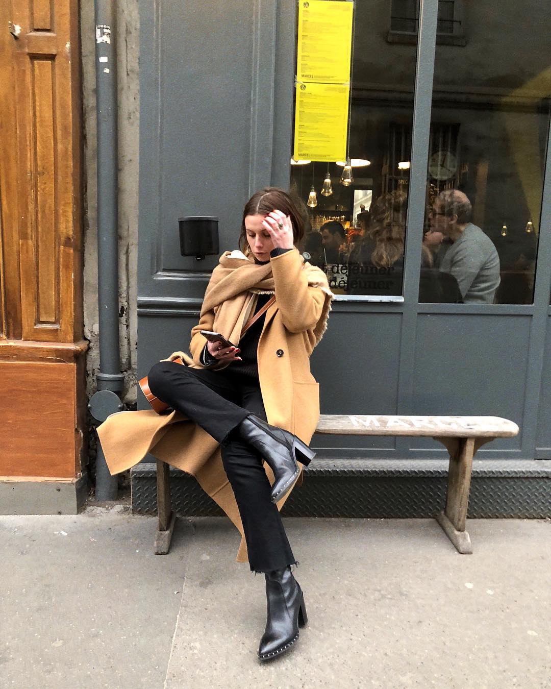 Stylish French-Girl Winter Outfit Idea — @solenelara Instagram look with a camel coat, small tan bucket bag, black raw-hem jeans, and black studded ankle boots