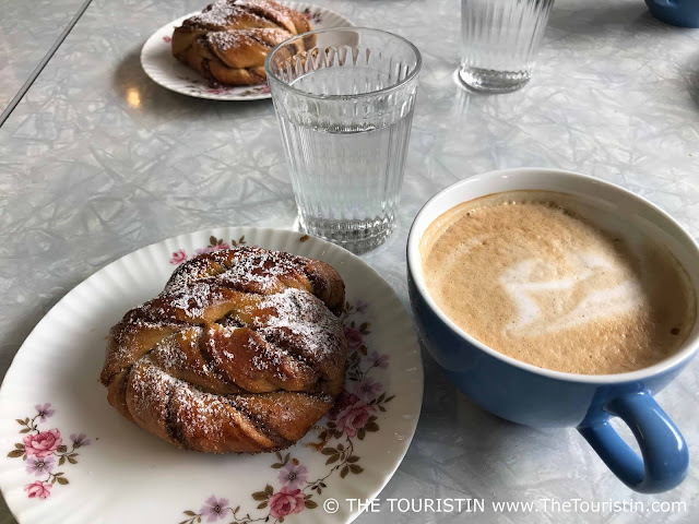 Two glasses of water, and a light blue china cup without a saucer filled with milked coffee next to two plates decorated with painted flower art serving icing sugar-coated danish, on a light grey table.