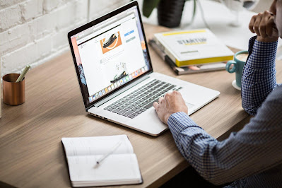 Male ads campaigner wearing a blue shirt working on computer, with notepads, brown cup, stack of books, a blue coffee cup on the working table.